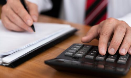 Pessoa realizando cálculos financeiros com calculadora e anotando em bloco de notas, representando planejamento tributário e impacto da reforma tributária.